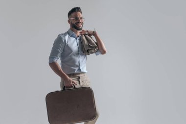 happy young man with jacket over shoulder holding suitcase and smiling while looking to side on grey background in studio