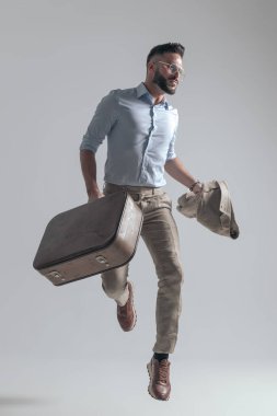 sexy bearded man looking to side while jumping in the air, holding suitcase and jacket on grey background in studio