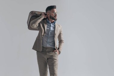 cool businessman with glasses holding hand in pocket and bag over shoulder while looking to side on grey background in studio