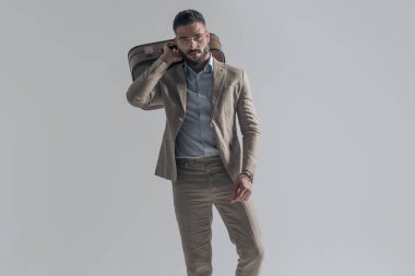 confident businessman with glasses holding briefcase over shoulder and posing in a fashion manner while walking on grey background in studio