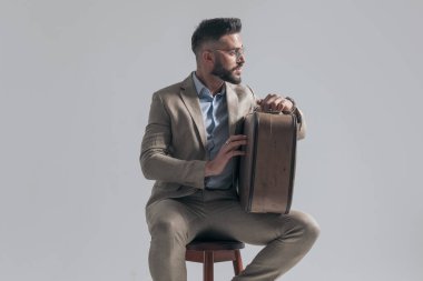 cool young model in suit with glasses looking to side and holding suitcase while sitting on grey background in studio