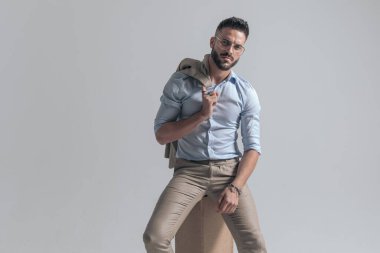 seductive bearded man with jacket over shoulder posing in a fashion way while sitting on grey background in studio