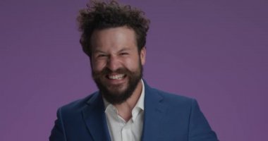 enthusiastic young guy in elegant suit making crazy faces, having fun, being idiot and enjoying, smiling and laughing in studio on blue background