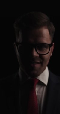 happy young businessman in suit wearing glasses, fixing tie and smiling on black background in a mysterious studio