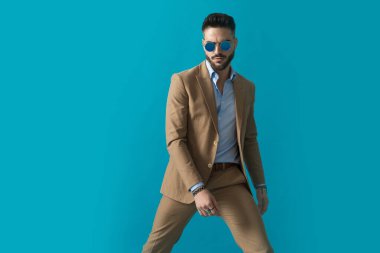 Bej takım elbiseli yakışıklı adam eski güneş gözlüğü takıyor. Modada kollarını tutuyor ve stüdyoda mavi arka planda poz veriyor.