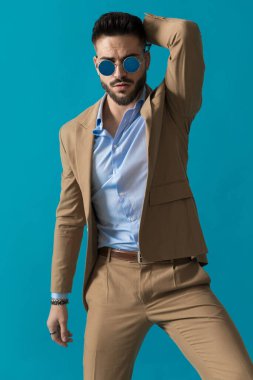 Takım elbiseli kendine güvenen bir işadamı boncuğun arkasında eski moda güneş gözlüğü takıyor ve stüdyoda mavi arka planda havalı duruyor.