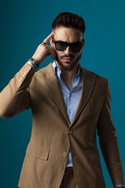 Havalı, traşsız, genç bir iş adamı. Parmakla işaret ediyor. Stüdyoda mavi arka planda gözlüklü bej bir takım elbiseyle yürüyor ve poz veriyor.