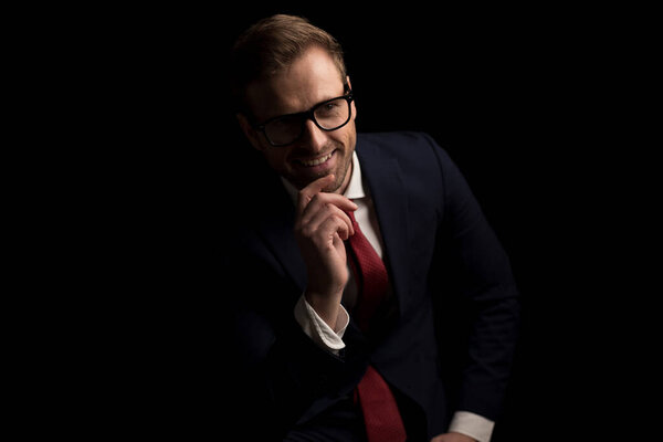 sexy businessman touching his chin while wearing eyeglasses, smiling and sitting on black background