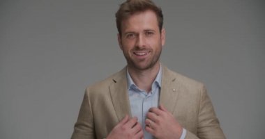 Yakışıklı iş adamı takım elbisesini düzeltiyor ve gri arka planda kameraya gülümsüyor.