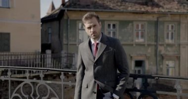 Zarif kıyafetli genç bir adam bavulu tutuyor, yan bakıyor, ortaçağın arka planında duruyor, gülümsüyor ve başka tarafa bakıyor.