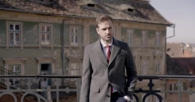 Genç işadamı çitlere yaslanıyor, bir elini cebinde ve çantasını tutuyor, paltosunu tamir ediyor.