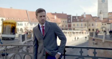 Zarif takım elbiseli kendine güvenen bir işadamı, uzun bir palto giyiyor, el ele tutuşuyor, yan yana bakıyor, kamera ortaçağ şehrinin dışında dururken gülümsüyor.