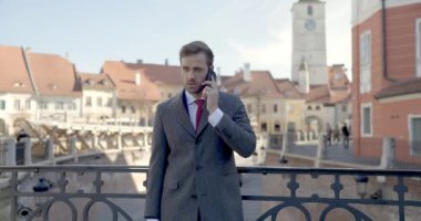 Bavullu seksi genç adam telefonda konuşuyor, güzel bir sohbet ediyor, ortaçağ kasabasının önünde dururken gülüyor ve gülümsüyor.