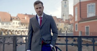 Cebinde uzun paltosu olan genç ve seksi bir işadamı, dışarıda bir köprüde duruyor, kamera yakınlaştığında gülümsüyor ve göz kırpıyor.