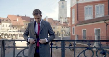 Ortaçağ kasabasının önünde uzun ceketini ilikleyen, baş parmak hareketi yapan ve dışarıda gülümseyen zarif, tıraşsız bir adam.