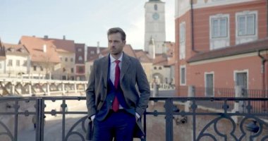 Yakışıklı iş adamı elleri ceplerinde çitlere yaslanıyor sonra ceketini kapatıyor ve gülümsüyor.