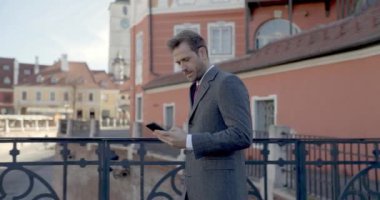 Çit boyunca yürüyen, telefonda konuşan ve iyi vakit geçiren yakışıklı bir iş adamının yan görüntüsü.