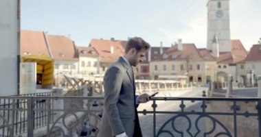 Zarif takım elbiseli, telefonu elinde tutan, Instagram 'da kayan, gülümseyen ve ortaçağ şehrinde yürürken arkasına bakan kendine güvenen bir adamın yan görüntüsü.