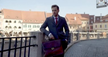 Yakışıklı iş adamı bir eli cebinde ve çantasında çitlere yaslanıyor sonra ceketini tamir ediyor ve gülümsüyor.