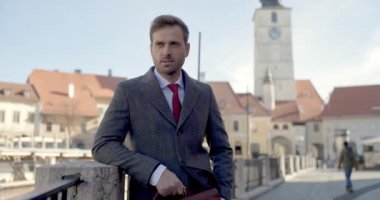 İş adamı çite yaslanıyor, bir eli cebinde, diğer eli çantasında başka tarafa bakıyor.