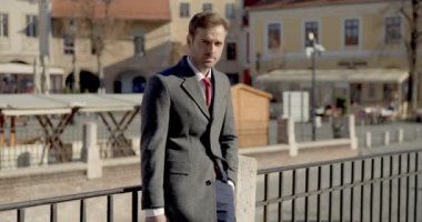 Çekici iş adamı çitlere yaslanıyor, bir evrak çantası ve elini cebinde tutuyor, başka tarafa bakıyor ve gülümsüyor.