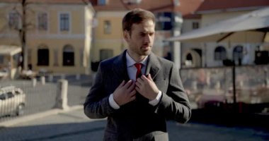 Takım elbiseli zarif bir genç adam uzun bir palto giyiyor, kenarda durup gülümsüyor.