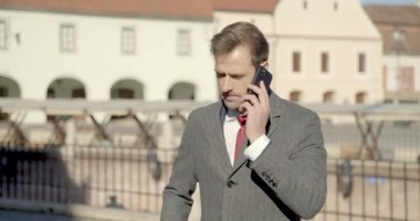 Takım elbiseli çekici iş adamı dışarıda telefonla konuşuyor ve ortaçağ arka planında dururken gülümsüyor.