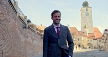 Sakallı, zarif iş adamı, el ele tutuşmuş, başka tarafa bakıyor ve gülümsüyor, dışarıda poz verirken ceketini düzenliyor.