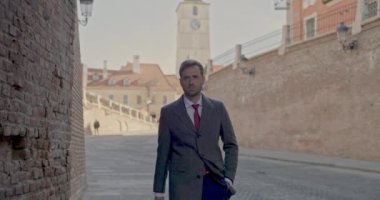 Seksi iş adamı bir eli cebinde ve elinde bir evrak çantasıyla duvarın yanında yürüyor ve ceketini tamir ediyor.