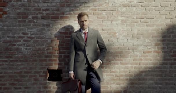 横を向いてシャツやネクタイやコートを調整し中世の壁の前で笑顔でスーツケースを持つセクシーな若い男背景 — ストック動画