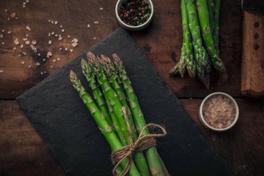 Tahta doğrama tahtasının üstünde çiğ kuşkonmaz demeti, eski ahşap bir masanın üstünde tuz ve biberle stil edilmiş. 