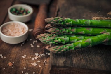 Taze kuşkonmazın yakın plan fotoğrafı pişmeye hazır, eski bir ahşap doğrama tahtasının üzerinde tuz, biber kaseleri ve klasik satırın yanında.