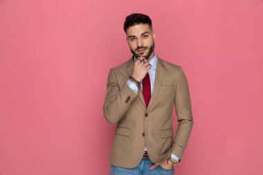 Genç yakışıklı işadamı, bir elini pembe arka planda cebine koyarak çenesine dokunuyor.