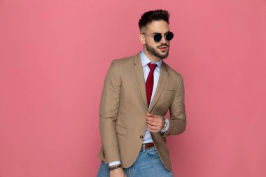 Yakışıklı iş adamı güneş gözlüğü takıyor, takım elbisesini düzenliyor ve pembe arka planda poz veriyor.