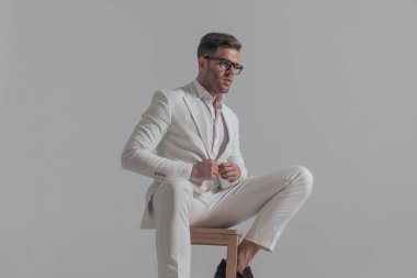 Yakışıklı iş adamı ceketini kapatırken bir sandalyede oturuyor ve beyaz arka plana bakıyor.