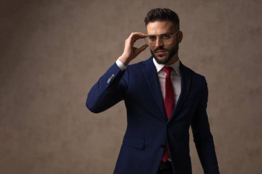Yakışıklı genç adam gözlüklerini düzeltiyor, güzel bir takım elbise giyiyor ve duvar kağıdına karşı poz veriyor.