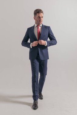 Stüdyodaki gri arka planda yürüyen ve kapanış ceketi giyen havalı genç iş adamının tam vücut fotoğrafı.