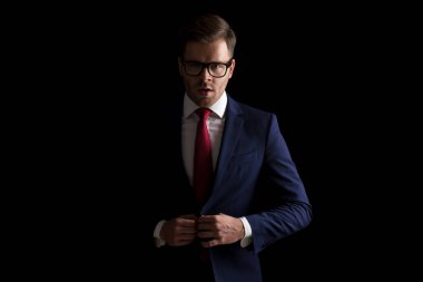 Moda ışığında takım elbise ayarlayan gözlüklü dramatik genç adam stüdyoda siyah arka plana karşı poz veriyor.