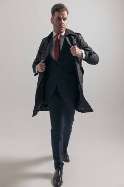 Zarif takım elbiseli seksi moda adamı stüdyoda gri arka planda yürürken uzun ceketini giyip ayarlamaya çalışıyor.