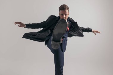 Takım elbiseli, uzun paltolu, bacağını kaldırıp stüdyoda gri arka planı tekmeleyen zarif bir genç.