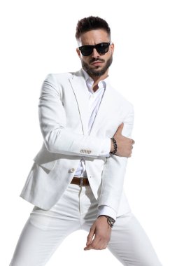 Tıraşsız ve güneş gözlüklü bir iş adamı beyaz arka planda moda stüdyosu ışığı altında havalı bir şekilde poz veriyor. 