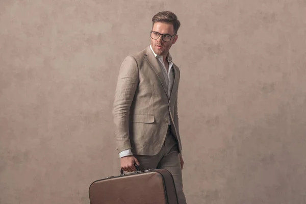 一位有魅力的商人 戴着眼镜 一边站在一边 一边提着手提箱 一边走在米色背景的工作室里 — 图库照片