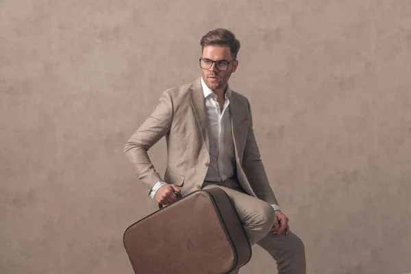 一个英俊的时尚男人拿着手提箱 坐在工作室米色的背景上 一边看一边看 — 图库照片