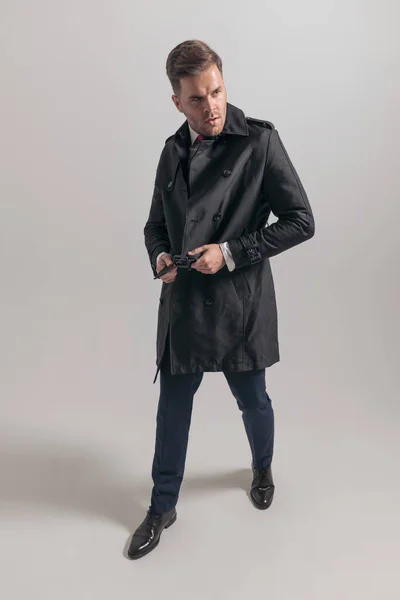 一位英俊的商人站在工作室的灰色背景上 一边看一边合上长大衣 — 图库照片