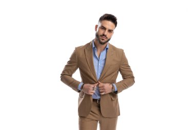 Beyaz arka planda stüdyoda poz verirken düğmelerini açıp kahverengi takım elbise giyen kendine güvenen genç adam.