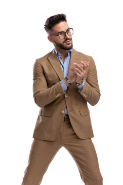 Kahverengi takım elbiseli havalı genç işadamı stüdyoda beyaz arka planda durup poz verirken avuçlarını ovalıyor ve yan tarafa bakıyor.