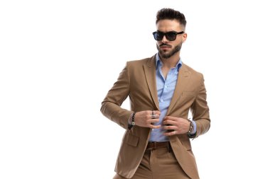 Güneş gözlüklü ve sakallı seksi esmer adam stüdyoda beyaz arka planda takım elbise giyiyor.