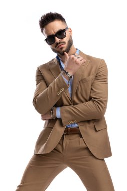 Güneş gözlüklü genç adam stüdyoda beyaz arka planda poz verirken kollarını kavuşturup çenesine dokunuyor.