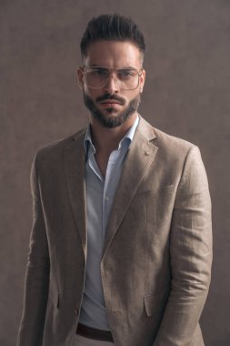 Gözlüklü sakallı genç işadamı stüdyoda kahverengi arka planda kendine güvenen bir şekilde poz veriyor.