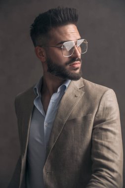Bej takım elbiseli yakışıklı sakallı bir adamın portresi. Gözlüğü yana bakıyor ve stüdyoda kahverengi arka planda poz veriyor.